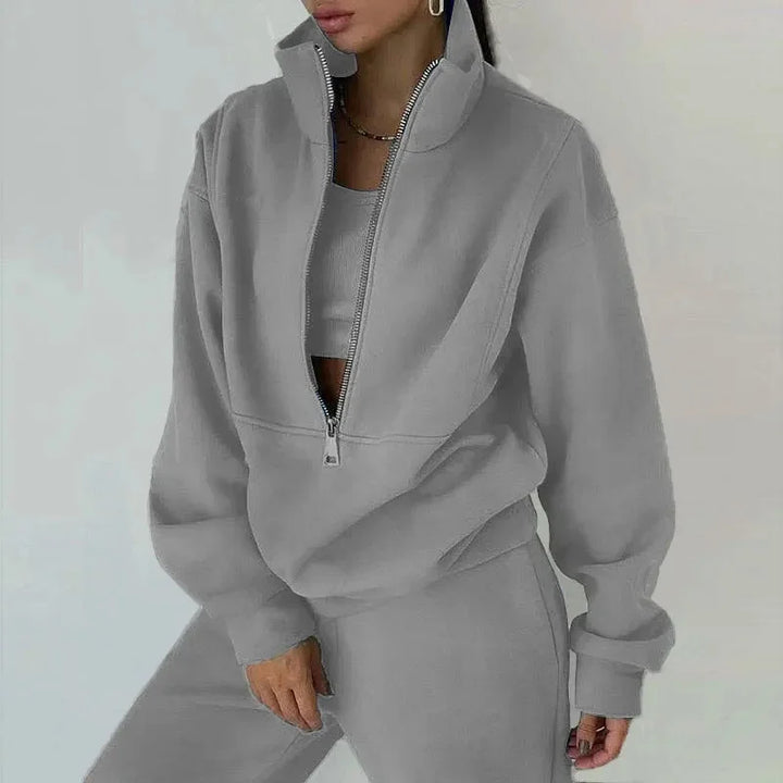 Une femme porte un ensemble décontracté gris clair composé d'un haut à zip et d'un pantalon assorti. Le haut est légèrement oversize avec un col zippé et elle a l'air détendue et élégante. Le fond est neutre et minimaliste.